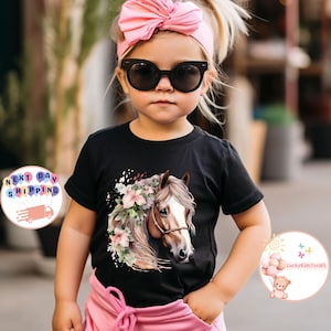 May include: A black t-shirt featuring a horse head design with floral accents. The horse has a brown bridle and is surrounded by pink and white flowers. The shirt is worn by a young child wearing sunglasses and a pink headband.