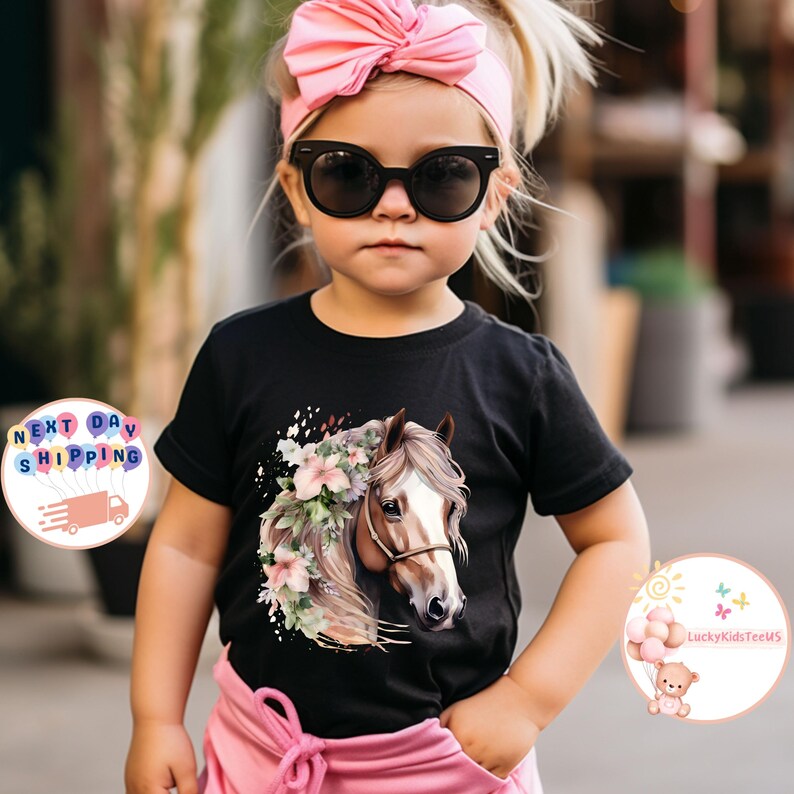 May include: A black t-shirt featuring a horse head design with floral accents. The horse has a brown bridle and is surrounded by pink and white flowers. The shirt is worn by a young child wearing sunglasses and a pink headband.