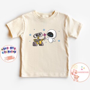 Puede incluir: Una camiseta de color crema con una imagen de dibujos animados de Wall-E y Eva de la pel&iacute;cula Wall-E. La imagen muestra a Wall-E sosteniendo un molinillo rosa y a Eva mir&aacute;ndolo con una sonrisa. Hay estrellas azules dispersas alrededor de la imagen.
