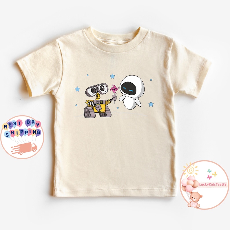 Puede incluir: Una camiseta de color crema con una imagen de dibujos animados de Wall-E y Eva de la pel&iacute;cula Wall-E. La imagen muestra a Wall-E sosteniendo un molinillo rosa y a Eva mir&aacute;ndolo con una sonrisa. Hay estrellas azules dispersas alrededor de la imagen.