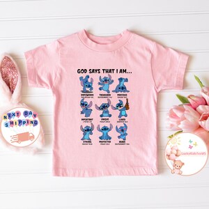 May include: Pink short-sleeved t-shirt with a blue cartoon character, Stitch, in various poses with bible verses. The text reads "God says that I am..." and lists words like "empowered", "treasured", "precious", "important", "unique", "loved", "strong", "protected", and "brave".