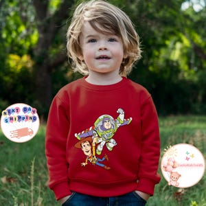 May include: A red sweatshirt featuring a graphic of Buzz Lightyear and Woody from Toy Story. The design is in bright colors and is centered on the front of the sweatshirt. The sweatshirt is worn by a young child.