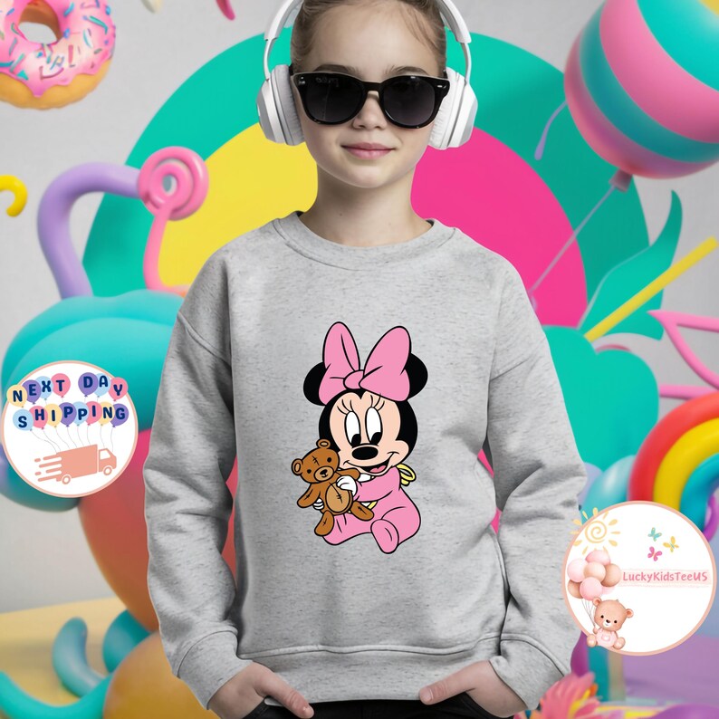 May include: A gray sweatshirt featuring a Minnie Mouse graphic. Minnie is depicted in a pink outfit, holding a teddy bear, and wearing a pink bow. The background includes colorful balloons and the text "Next Day Shipping".