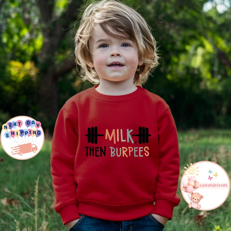 May include: A red sweatshirt with the text "MILK THEN BURPEES" in a colorful font and a barbell graphic. The sweatshirt is worn by a young child with blonde hair, standing in front of a blurred green background.