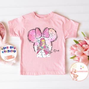 May include: Pink t-shirt featuring a cartoon illustration of Bo Peep from Toy Story. Bo Peep is wearing a blue dress with white polka dots and a pink bow. She is holding a shepherd's crook and standing in front of a pink castle. The image is surrounded by a pink and white Mickey Mouse ear silhouette. The text "Bo Peep" is written in pink below the image.