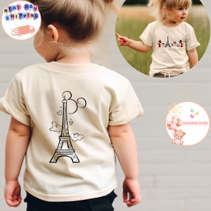 May include: A beige short-sleeved t-shirt with a black and white illustration of the Eiffel Tower and Mickey Mouse ears on the back. The front of the shirt has a black and white illustration of Mickey Mouse, Minnie Mouse, Donald Duck, and Goofy in front of the Eiffel Tower with the word "Paris" written below.