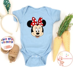 May include: A pale blue baby bodysuit with a Minnie Mouse design, including a red polka dot bow. The image includes baby shoes, a bunny ear headband, and decorative carrots. The text "Next Day Shipping" is also visible.