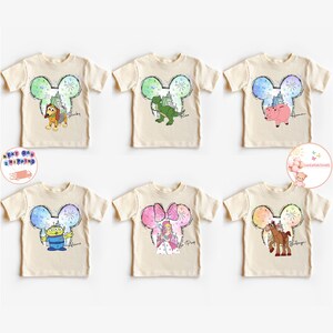 May include: Six cream coloured t-shirts with a Mickey Mouse ear design featuring different Disney characters. The characters include Woody, Rex, Hamm, Bo Peep, Bullseye, and a pig. Each shirt has the character's name printed below the image.
