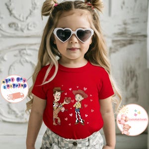 May include: A young child wearing a red t-shirt featuring a cartoon image of Woody and Jessie from Toy Story, with heart-shaped accents. The child is also wearing heart-shaped sunglasses.