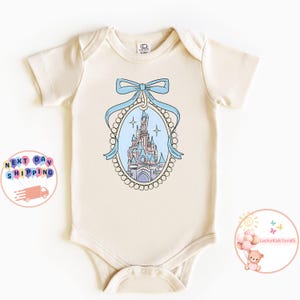 Puede incluir: Un body de beb&eacute; de color crema con una ilustraci&oacute;n azul y rosa de un castillo con un lazo azul en la parte superior. El castillo est&aacute; rodeado por un borde de perlas.
