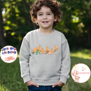May include: A light gray sweatshirt featuring a colorful graphic of cartoon characters with orange traffic cones. The design includes characters in various colors. The sweatshirt is worn by a child.