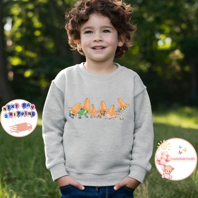 May include: A light gray sweatshirt featuring a colorful graphic of cartoon characters with orange traffic cones. The design includes characters in various colors. The sweatshirt is worn by a child.