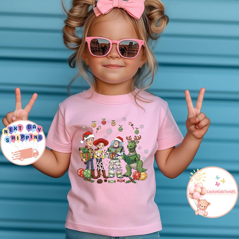 May include: A pink t-shirt with a festive Toy Story graphic featuring Woody, Jessie, Buzz Lightyear, and Rex in Christmas outfits. The image also shows a child wearing pink sunglasses and a pink bow.