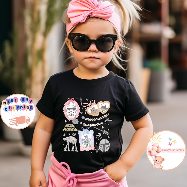 May include: A child's black t-shirt featuring a Star Wars design with pink and white accents. The graphic includes characters and symbols from the franchise. The child is wearing sunglasses and a pink headband.