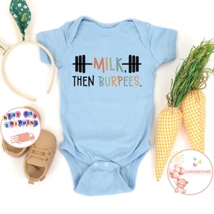 May include: A light blue baby onesie with the text "MILK THEN BURPEES" in black, orange, and green. The onesie has a barbell graphic. The image also includes a pair of baby shoes, a bunny ear headband, and decorative carrots.