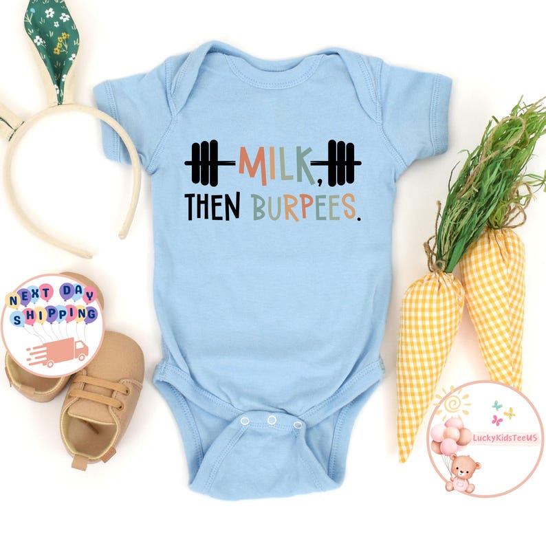 May include: A light blue baby onesie with the text "MILK THEN BURPEES" in black, orange, and green. The onesie has a barbell graphic. The image also includes a pair of baby shoes, a bunny ear headband, and decorative carrots.