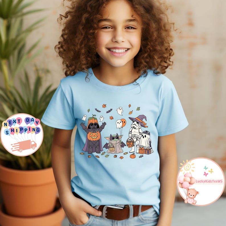 以下が含まれることがあります： ハロウィーンをテーマにしたデザインのライトブルーのTシャツ。デザインには、カボチャの頭をしたダース・ベイダー、ベビーヨーダ、ストームトルーパー、幽霊が含まれています。シャツは茶色のベルトで着用されています。