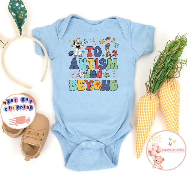Puede incluir: Un body de beb&eacute; azul claro con un gr&aacute;fico de Woody y Buzz Lightyear de Toy Story. El gr&aacute;fico dice "To Autism and Beyond" con piezas de rompecabezas y estrellas.