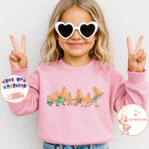 May include: A pink sweatshirt featuring a cartoon design of characters wearing orange hats. The design includes a green dinosaur, a brown dog, a pink pig, and a blue and white character. The child is wearing white heart-shaped sunglasses and making a peace sign.