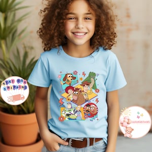 Op de afbeelding: Een lichtblauw T-shirt met een grafische print van personages uit de Toy Story-filmfranchise. De personages zijn in een kleurrijk geometrisch ontwerp met een retro-esthetiek uit de jaren 90.