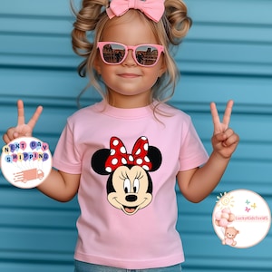 May include: A light pink t-shirt with a Minnie Mouse graphic, featuring a red and white polka dot bow. The shirt is worn by a child wearing pink sunglasses and a pink bow. The background is a blue wall.