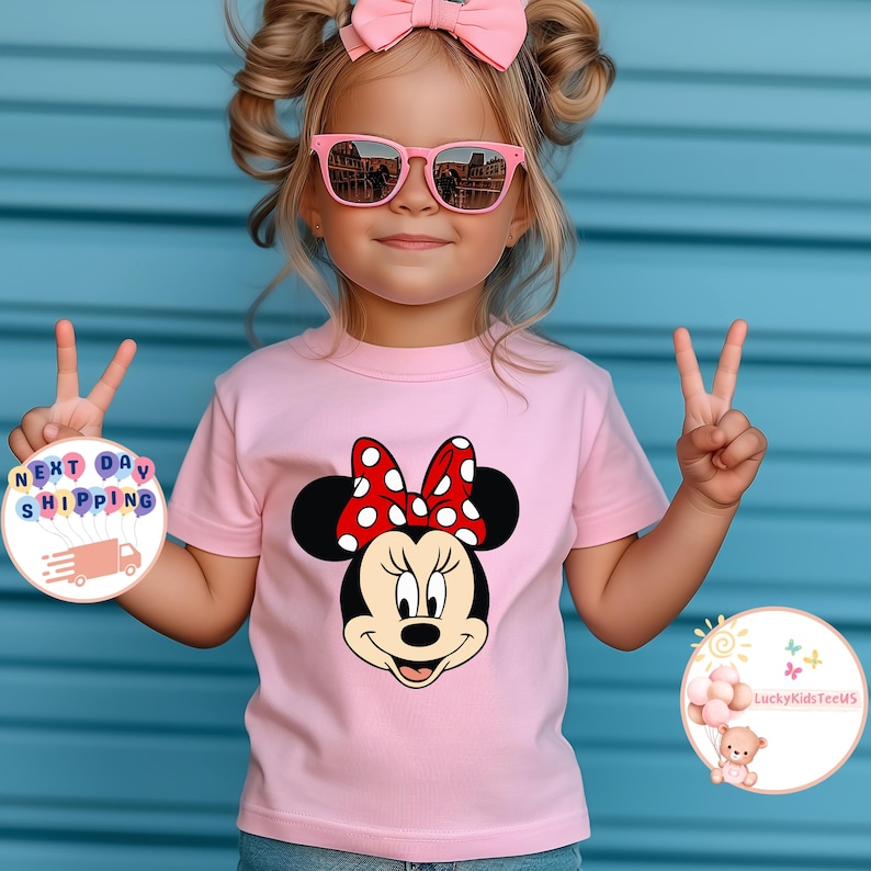 May include: A light pink t-shirt with a Minnie Mouse graphic, featuring a red and white polka dot bow. The shirt is worn by a child wearing pink sunglasses and a pink bow. The background is a blue wall.