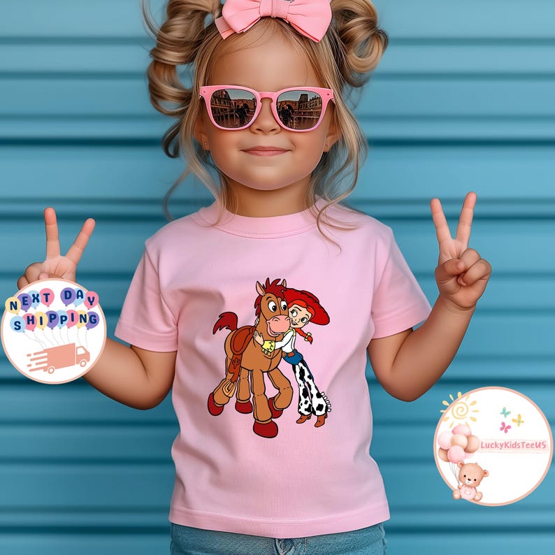 May include: A light pink t-shirt featuring a cartoon graphic of a horse and a cowgirl hugging. The shirt is worn by a young child with pink sunglasses and a pink bow. The background is a blue wall.