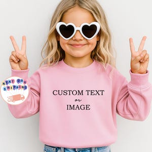 May include: A light pink sweatshirt with the text "CUSTOM TEXT or IMAGE" in black. The person in the image is wearing white heart-shaped sunglasses and making a peace sign with their fingers.