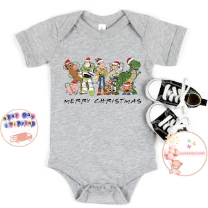 May include: A gray baby onesie with a Christmas-themed design featuring characters from the Toy Story franchise. The design includes Woody, Buzz Lightyear, Jessie, and other characters. The text "Merry Christmas" is printed below the characters.