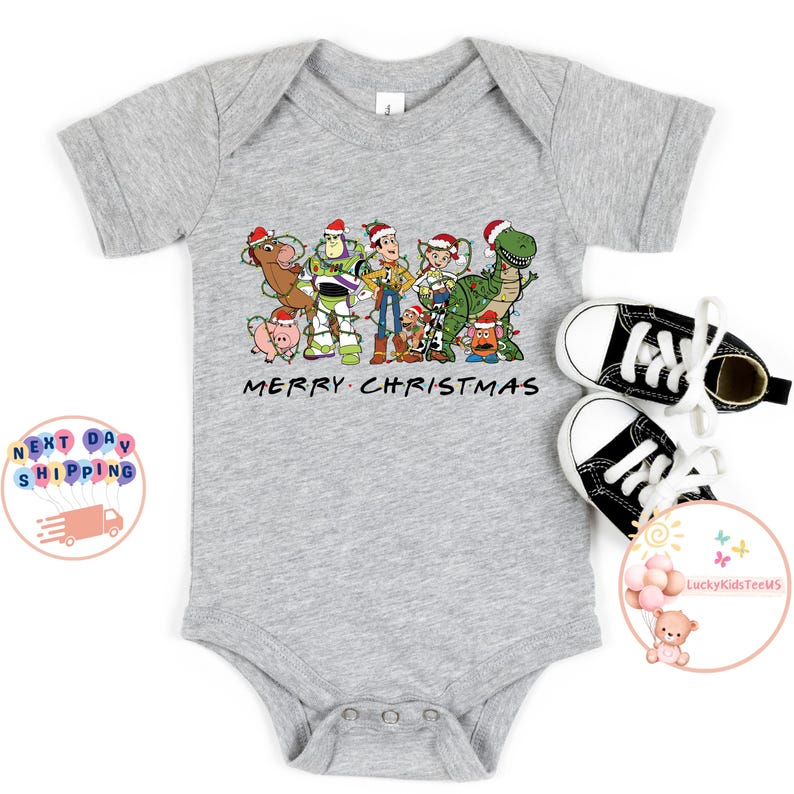 May include: A gray baby onesie with a Christmas-themed design featuring characters from the Toy Story franchise. The design includes Woody, Buzz Lightyear, Jessie, and other characters. The text "Merry Christmas" is printed below the characters.