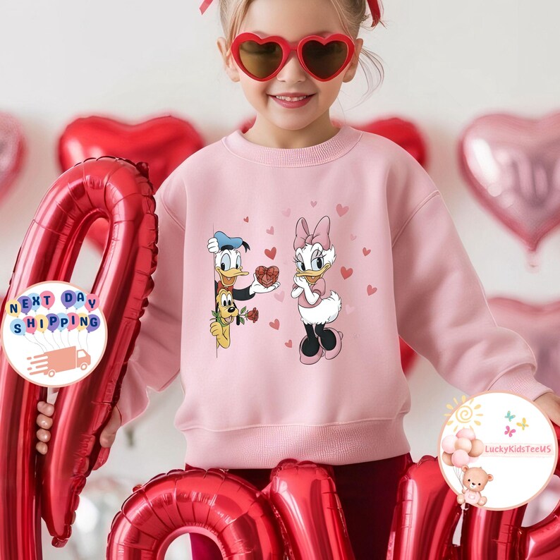 May include: A light pink sweatshirt featuring Donald Duck and Daisy Duck with hearts. Donald Duck is holding a heart-shaped box of chocolates, and Daisy Duck is looking at him. The sweatshirt has a crew neck and long sleeves. The background has red heart-shaped balloons.