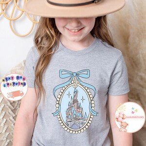Puede incluir: Una camiseta gris claro con una ilustraci&oacute;n azul y blanca de un castillo en un marco incrustado de perlas con un lazo azul en la parte superior. El castillo tiene detalles en rosa y marr&oacute;n.