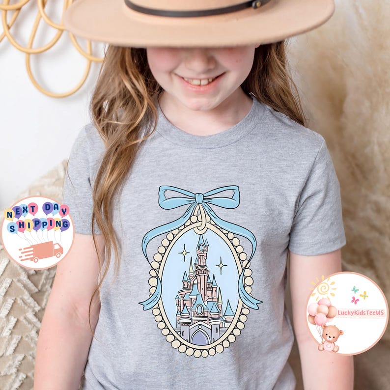 Puede incluir: Una camiseta gris claro con una ilustraci&oacute;n azul y blanca de un castillo en un marco incrustado de perlas con un lazo azul en la parte superior. El castillo tiene detalles en rosa y marr&oacute;n.