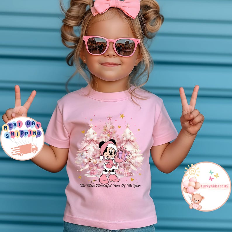 May include: A pink t-shirt featuring a Minnie Mouse graphic holding a gift in front of Christmas trees. The text "The Most Wonderful Time Of The Year" is below the graphic. The child is wearing pink sunglasses and a pink bow.