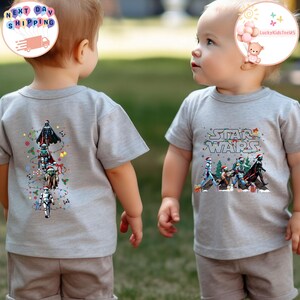 May include: Two children wearing grey t-shirts. One shirt has a vertical design of Star Wars characters, the other displays the Star Wars logo with characters. Both shirts feature festive holiday elements.