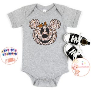 May include: A grey baby onesie with a floral pumpkin design in the shape of Mickey Mouse's head. The pumpkin has a brown stem and a smiling face.
