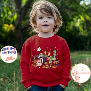 Puede incluir: Una sudadera roja con un dise&ntilde;o de Halloween de personajes de Toy Story. El dise&ntilde;o incluye a Woody, Buzz Lightyear y otros personajes, con calabazas, fantasmas y murci&eacute;lagos. La sudadera la lleva un ni&ntilde;o peque&ntilde;o.