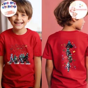 May include: Red t-shirt with Star Wars characters and text. The front design shows the Star Wars logo and characters with Christmas tree decorations. The back of the shirt displays Darth Vader, a Stormtrooper, and Yoda.
