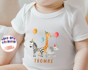 May include: White baby onesie featuring a colorful illustration of a zebra, giraffe, and elephant, each wearing a party hat and holding a balloon. The name "THOMAS" is printed in orange below the animals. A cheerful design for a baby.