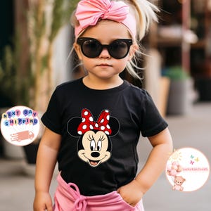 May include: A black t-shirt with a Minnie Mouse design, showcasing a red bow with white polka dots. The image shows a child wearing the tee, complemented by a pink headband and sunglasses. This is a children's clothing item.