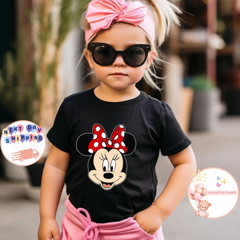 May include: A black t-shirt with a Minnie Mouse design, showcasing a red bow with white polka dots. The image shows a child wearing the tee, complemented by a pink headband and sunglasses. This is a children's clothing item.