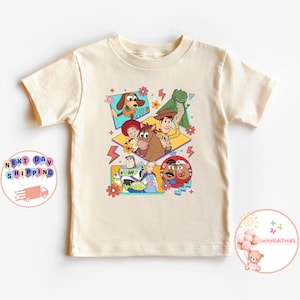 Op de afbeelding: Een beige T-shirt met een kleurrijke grafische print van personages uit de Toy Story-filmfranchise. De personages zijn Woody, Buzz Lightyear, Jessie, Rex en Slinky Dog. De grafiek is in een retro 90s-stijl met felle kleuren en geometrische vormen.