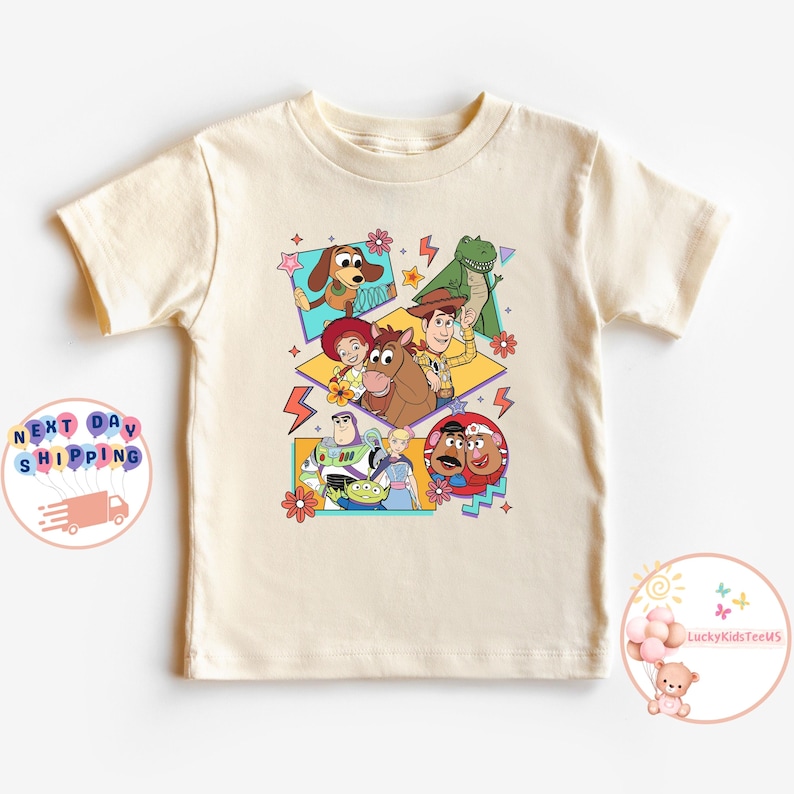 Op de afbeelding: Een beige T-shirt met een kleurrijke grafische print van personages uit de Toy Story-filmfranchise. De personages zijn Woody, Buzz Lightyear, Jessie, Rex en Slinky Dog. De grafiek is in een retro 90s-stijl met felle kleuren en geometrische vormen.