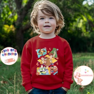 Op de afbeelding: Rode longsleeve trui met een kleurrijke grafische print van personages uit de Toy Story-filmfranchise. De personages zijn Woody, Buzz Lightyear, Jessie, Rex en Mr. Potato Head.