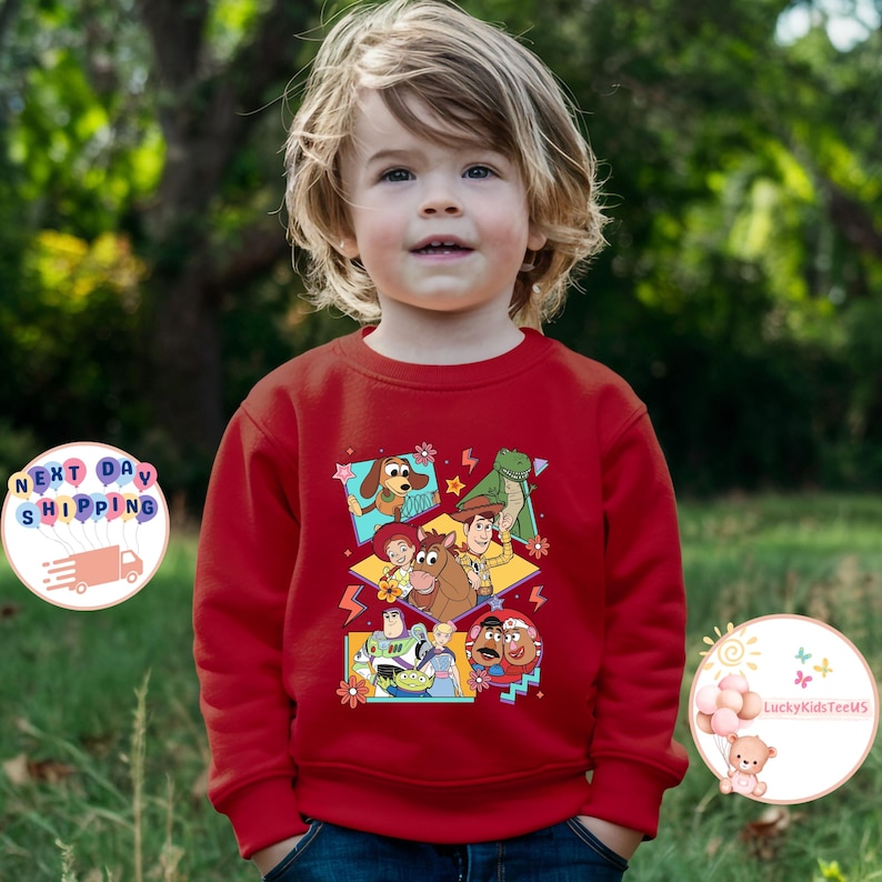 Op de afbeelding: Rode longsleeve trui met een kleurrijke grafische print van personages uit de Toy Story-filmfranchise. De personages zijn Woody, Buzz Lightyear, Jessie, Rex en Mr. Potato Head.
