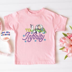 May include: A light pink t-shirt featuring a graphic of Buzz Lightyear from Toy Story. The design includes the phrase "To Infinity" in green and the word "Infinity" in purple script with blue stars.