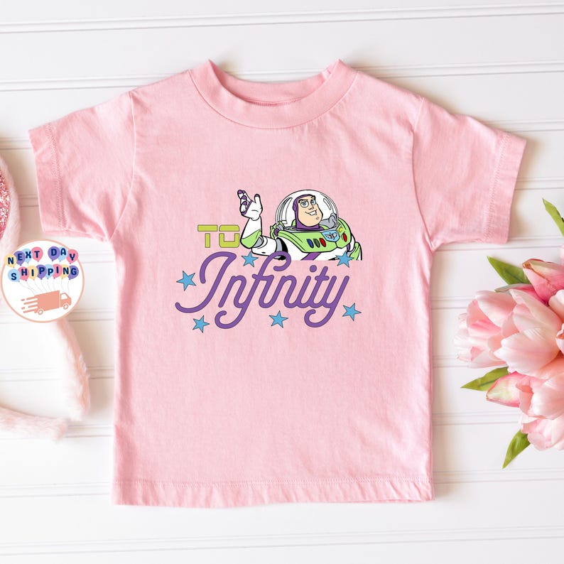 May include: A light pink t-shirt featuring a graphic of Buzz Lightyear from Toy Story. The design includes the phrase "To Infinity" in green and the word "Infinity" in purple script with blue stars.