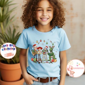 May include: A light blue t-shirt with a Christmas-themed Toy Story graphic. Woody, Jessie, Buzz Lightyear, and Rex are shown wearing Santa hats and festive decorations. The t-shirt is worn by a child.