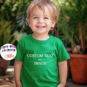 May include: A smiling child wearing a green t-shirt with the words "CUSTOM TEXT or IMAGE" in white. The child is in front of a blurred background of green plants and a brown pot.