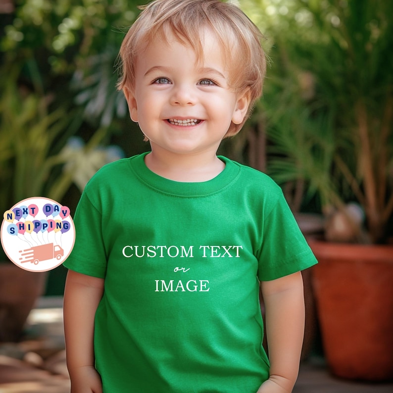 May include: A smiling child wearing a green t-shirt with the words "CUSTOM TEXT or IMAGE" in white. The child is in front of a blurred background of green plants and a brown pot.